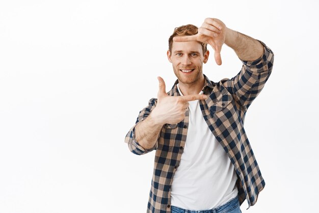 Souriant beau mec rousse faisant des cadres de main geste de l'appareil photo et prendre une photo dans son instant de mémoire à la recherche de plaisir debout sur fond blanc