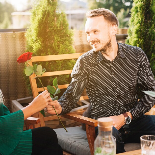 Souriant bel homme assis sur une chaise donnant rose rouge à sa petite amie