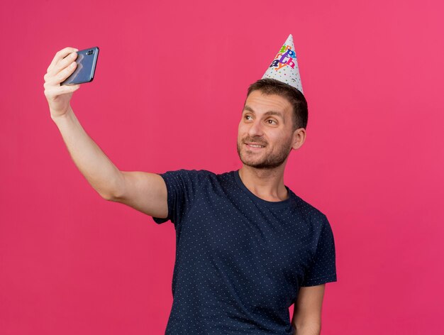 Souriant bel homme caucasien portant une casquette d'anniversaire tient et regarde le téléphone prenant selfie isolé sur fond rose avec espace de copie