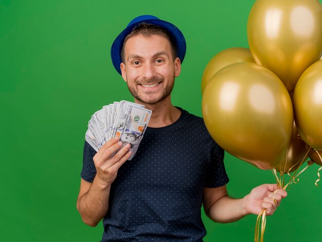 Souriant bel homme caucasien portant chapeau de fête bleu détient de l'argent et des ballons d'hélium isolés sur fond vert avec espace de copie