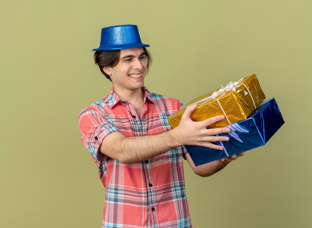 Souriant bel homme caucasien portant un chapeau de fête bleu tient et regarde les coffrets cadeaux