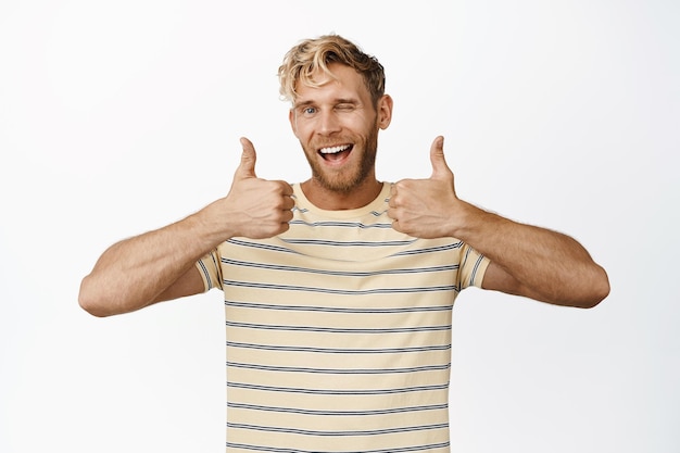 Souriant bel homme montrant les pouces vers le haut et un clin d'œil dire oui louange et compliment debout en t-shirt sur fond blanc