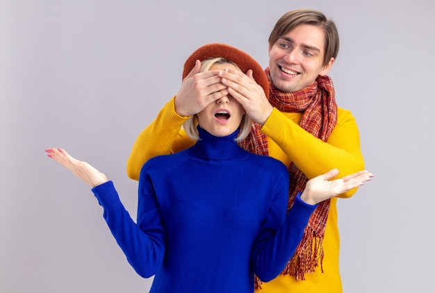 Souriant Bel Homme Slave Avec Une écharpe Autour Du Cou Fermant Les Yeux D'une Jolie Femme Blonde Surprise Avec Un Béret Isolé Sur Un Mur Blanc Avec Espace De Copie