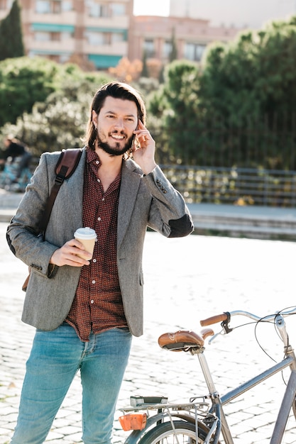 Souriant bel homme tenant une tasse de café à emporter parler sur téléphone portable dans le parc