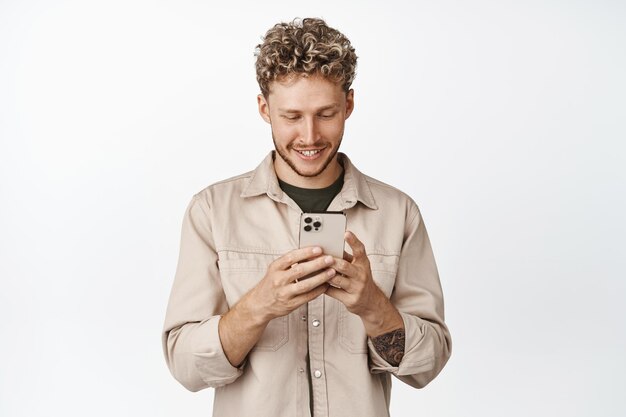 Souriant caucasien gars à l'aide de smartphone bel homme chat sur téléphone portable debout sur fond blanc