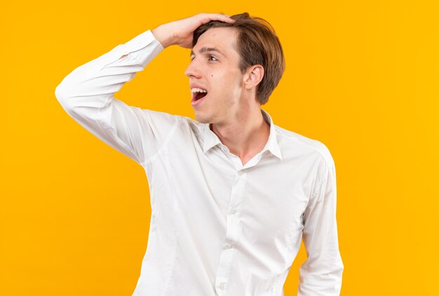 Souriant à côté d'un jeune beau mec vêtu d'une chemise blanche mettant la main sur la tête isolée sur un mur orange