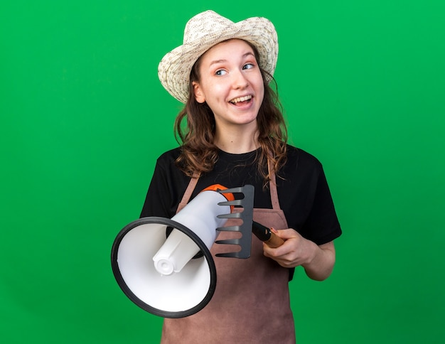 Souriant à côté jeune femme jardinier portant chapeau de jardinage tenant haut-parleur avec râteau