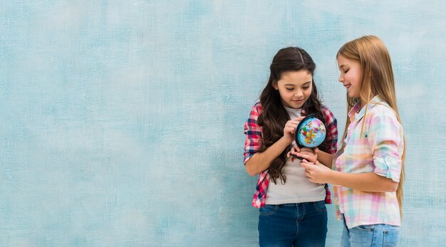 Souriant deux filles debout contre le mur bleu à la recherche d&#39;un petit globe