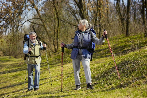 Souriant et heureux ensemble. Couple de famille âgés d'homme et de femme en tenue de touriste marchant sur la pelouse verte en journée ensoleillée près du ruisseau. Concept de tourisme, mode de vie sain, détente et convivialité.