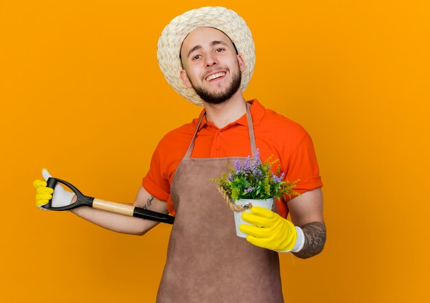 Souriant jardinier mâle portant chapeau et gants de jardinage détient des fleurs en pot de fleurs et pelle derrière le dos