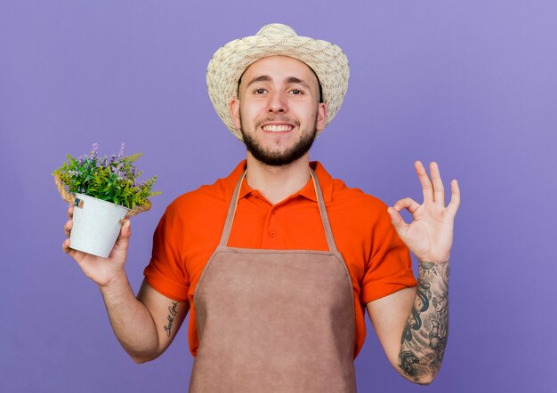 Souriant jardinier mâle portant des gestes de chapeau de jardinage signe de la main ok et détient pot de fleurs