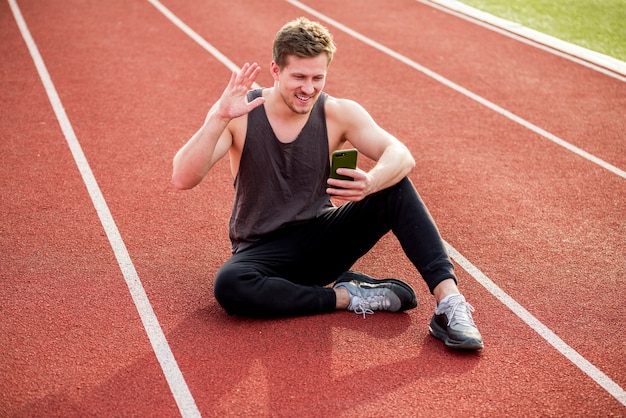 Souriant jeune athlète masculin assis sur une piste de course rouge agitant ses mains tout en faisant un appel vidéo