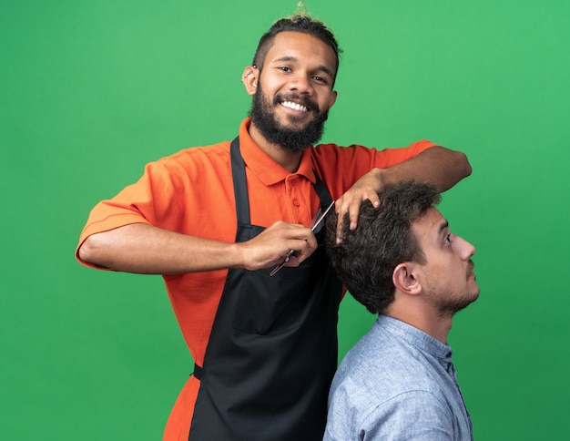 Souriant jeune barbier afro-américain en uniforme faisant une coupe de cheveux pour son jeune client