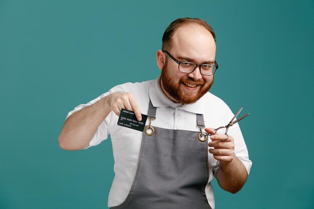 Souriant jeune barbier en uniforme et lunettes regardant la caméra montrant la carte de crédit et les ciseaux isolés sur fond bleu