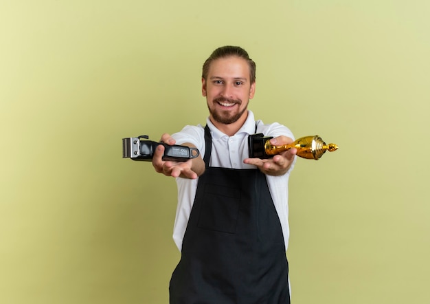 Photo gratuite souriant jeune beau coiffeur étirement tondeuse à cheveux et coupe gagnant vers l'avant isolé sur mur vert olive