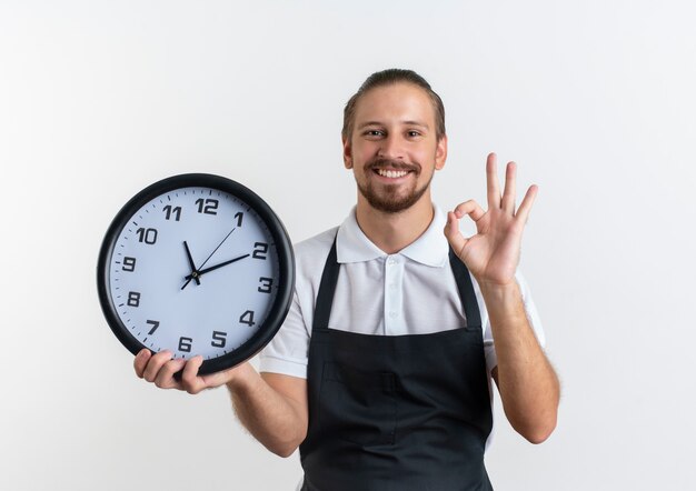 Souriant jeune beau coiffeur en uniforme tenant horloge et faisant signe ok isolé sur mur blanc