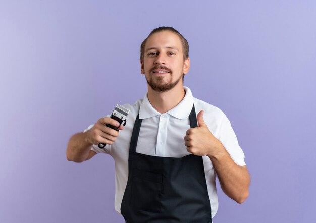 Souriant jeune beau coiffeur en uniforme tenant une tondeuse à cheveux et montrant le pouce vers le haut isolé sur un mur violet