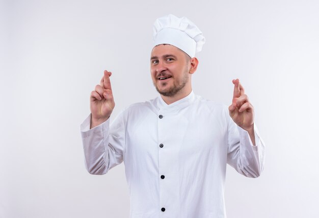 Souriant jeune beau cuisinier en uniforme de chef faisant le geste des doigts croisés isolé sur l'espace blanc