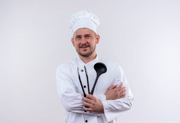 Souriant jeune beau cuisinier en uniforme de chef tenant une louche et une pince à griller isolé sur un espace blanc