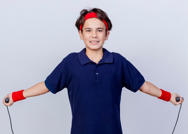 Souriant jeune beau garçon sportif portant un bandeau et des bracelets avec un appareil dentaire en regardant la caméra à la corde à sauter isolé sur fond blanc