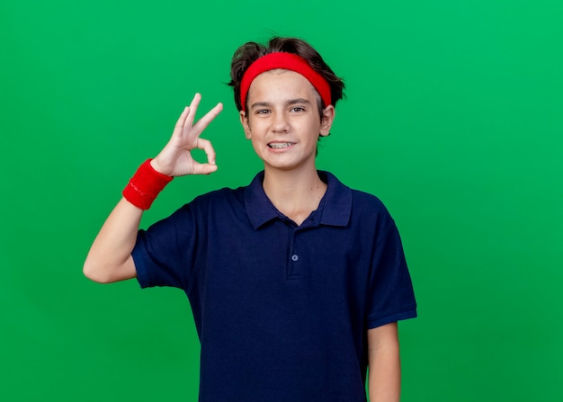 Souriant jeune beau garçon sportif portant bandeau et bracelets avec appareil dentaire regardant la caméra faisant signe ok isolé sur fond vert avec espace copie