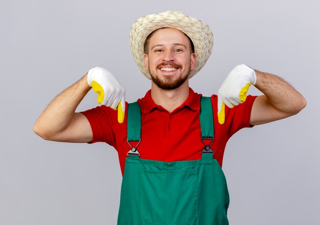 Souriant jeune beau jardinier slave en uniforme portant des gants de jardinage et un chapeau pointant vers le bas isolé sur un mur blanc