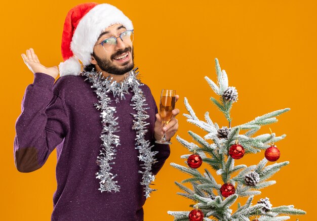 Souriant jeune beau mec debout à proximité de l'arbre de Noël portant un chapeau de Noël avec guirlande sur le cou tenant un verre de champagne isolé sur fond orange