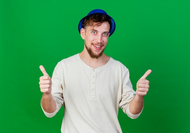 Souriant jeune beau mec de fête slave portant chapeau de fête regardant la caméra montrant les pouces vers le haut isolé sur fond vert