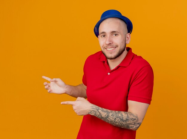 Souriant jeune beau mec de fête slave portant chapeau de fête regardant la caméra pointant sur le côté isolé sur fond orange avec espace de copie