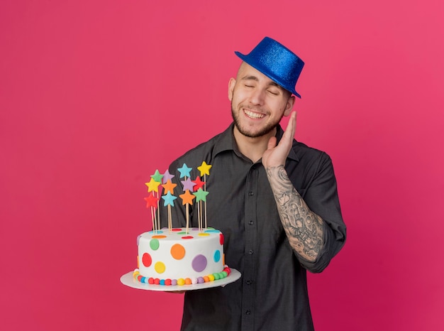 Souriant jeune beau mec de fête slave portant chapeau de fête tenant le gâteau d'anniversaire avec des étoiles gardant la main dans l'air avec les yeux fermés isolé sur fond cramoisi avec espace copie