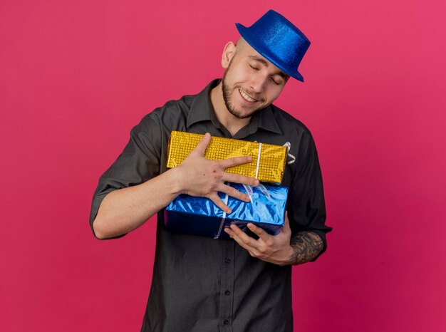 Souriant jeune beau mec de fête slave portant chapeau de fête tenant des paquets-cadeaux avec les yeux fermés isolés sur fond cramoisi avec espace copie