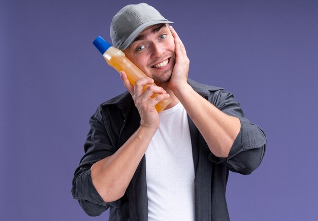 Souriant jeune beau mec de nettoyage portant un t-shirt et une casquette tenant un agent de nettoyage sur la joue isolé sur un mur violet