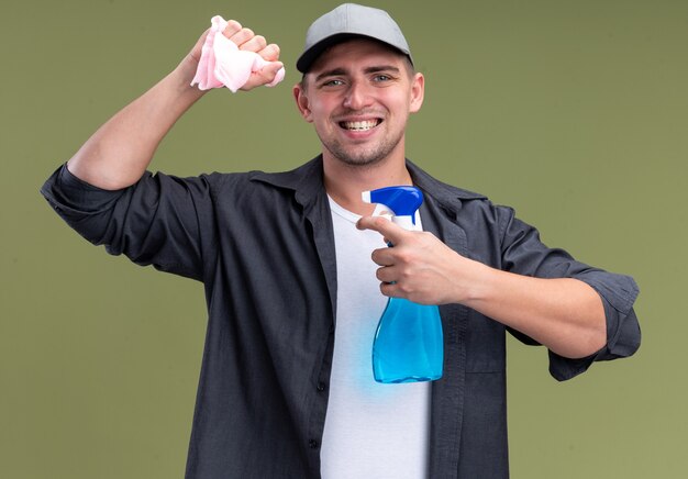 Photo gratuite souriant jeune beau mec de nettoyage portant un t-shirt et une casquette tenant un chiffon avec un vaporisateur isolé sur un mur vert olive