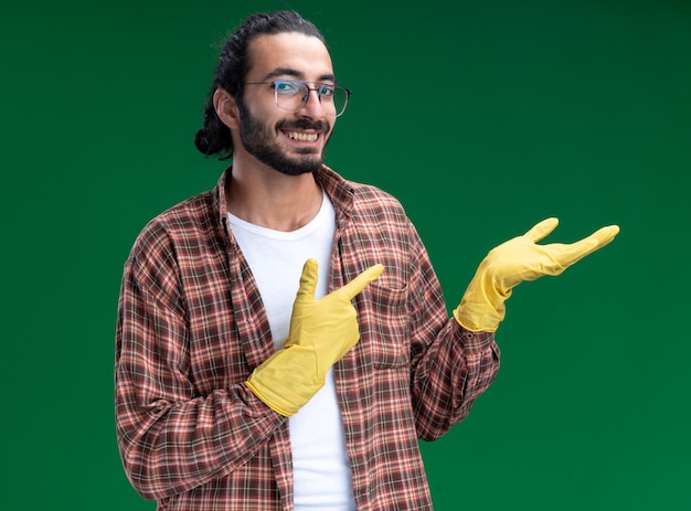 Souriant jeune beau mec de nettoyage portant un t-shirt et des gants faisant semblant de tenir et pointe quelque chose d'isolé sur le mur vert avec espace copie