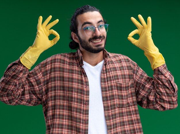 Souriant jeune beau mec de nettoyage portant un t-shirt et des gants montrant un geste correct isolé sur un mur vert