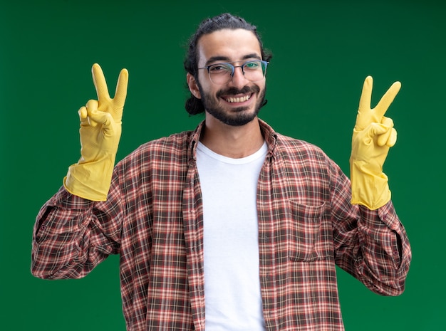 Souriant jeune beau mec de nettoyage portant un t-shirt et des gants montrant un geste de paix isolé sur un mur vert