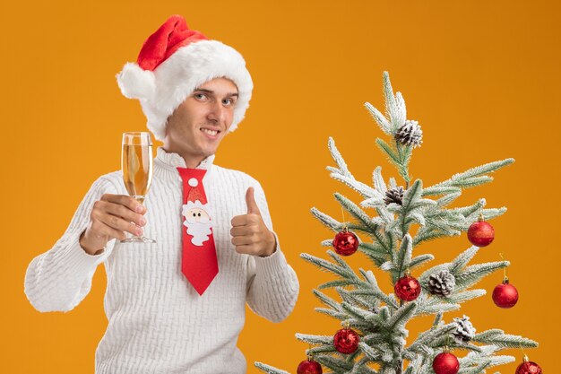 Souriant jeune beau mec portant chapeau de Noël et cravate de père Noël debout près de sapin de Noël décoré tenant un verre de champagne regardant la caméra montrant le pouce vers le haut isolé sur fond orange