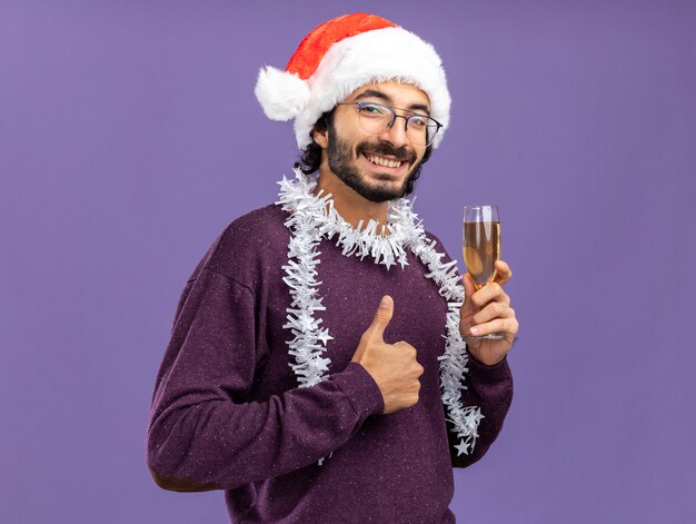 Souriant jeune beau mec portant un chapeau de Noël avec guirlande sur le cou tenant un verre de champagne montrant le pouce vers le haut isolé sur fond bleu
