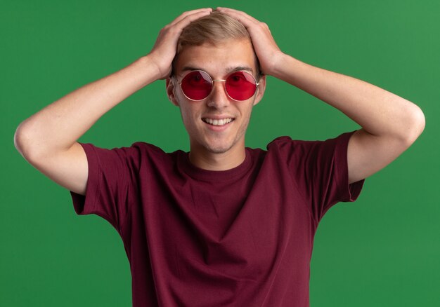 Souriant jeune beau mec portant une chemise rouge et des lunettes a attrapé la tête isolée sur le mur vert