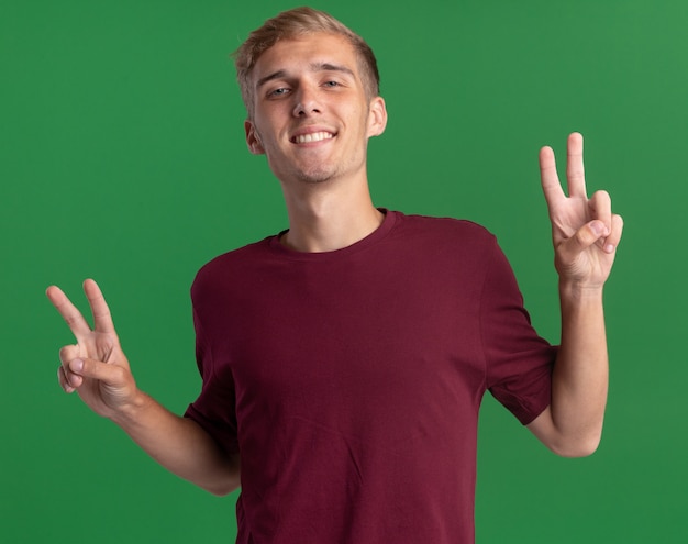 Souriant jeune beau mec portant une chemise rouge montrant le geste de paix isolé sur le mur vert