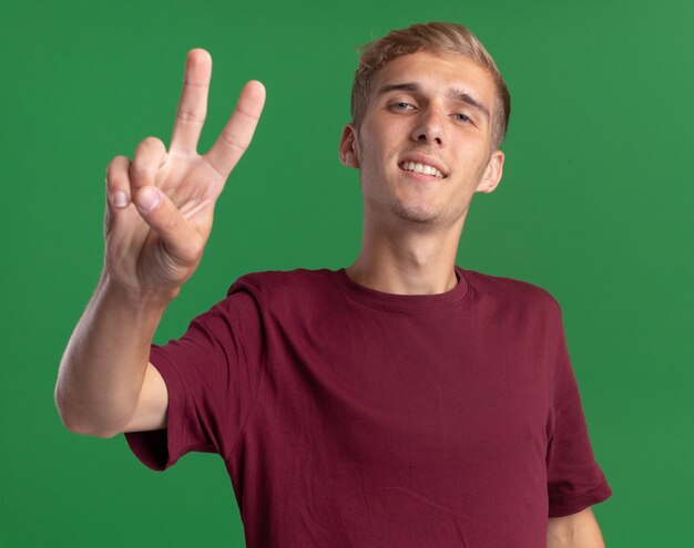 Souriant jeune beau mec portant une chemise rouge montrant le geste de paix isolé sur le mur vert