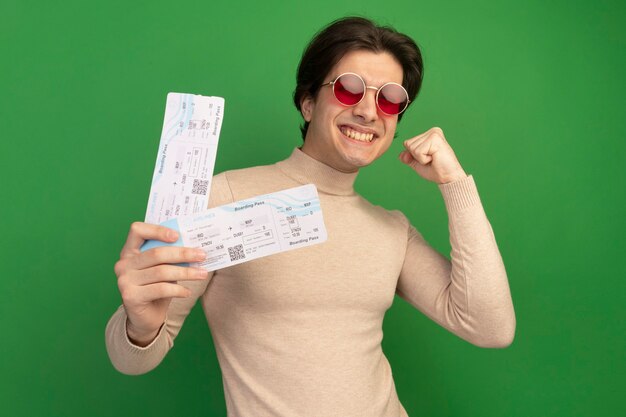 Souriant jeune beau mec portant des lunettes tenant des billets montrant oui geste isolé sur mur vert