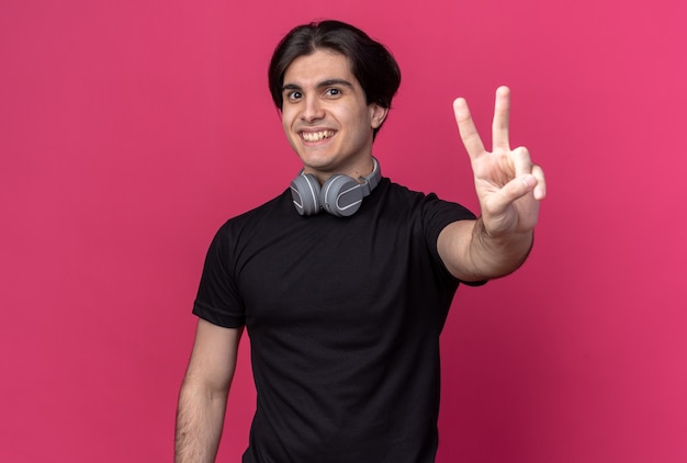 Souriant jeune beau mec portant un t-shirt noir et des écouteurs autour du cou montrant le geste de paix isolé sur un mur rose