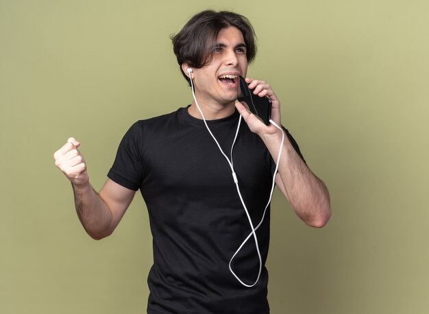 Souriant jeune beau mec portant un t-shirt noir avec écouteurs tenant le téléphone et chante isolé sur mur vert olive