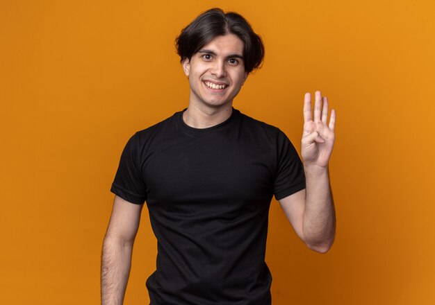 Souriant jeune beau mec portant un t-shirt noir montrant quatre isolés sur un mur orange