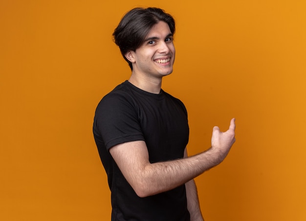 Souriant jeune beau mec portant un t-shirt noir pointe derrière isolé sur un mur orange avec espace pour copie