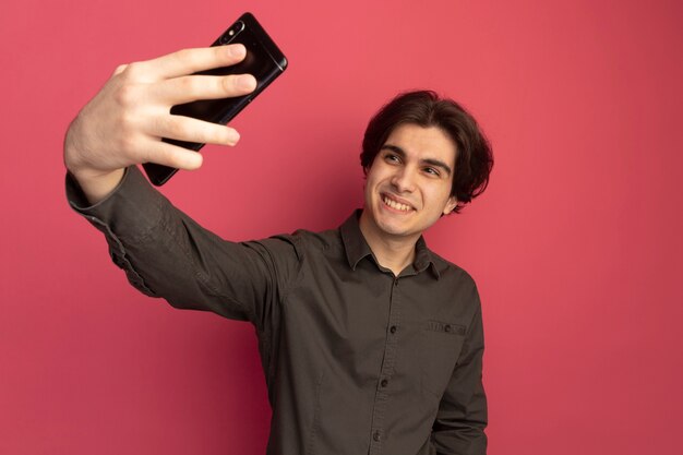 Souriant jeune beau mec portant un t-shirt noir prendre un selfie isolé sur un mur rose