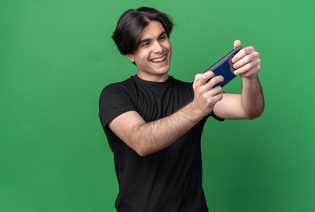 Souriant jeune beau mec portant un t-shirt noir prendre un selfie isolé sur un mur vert avec espace copie