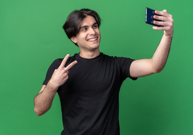 Souriant jeune beau mec portant un t-shirt noir prendre un selfie montrant le geste de paix isolé sur le mur vert