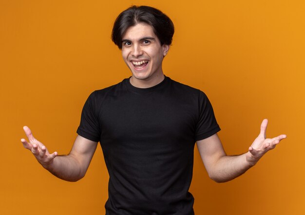 Souriant jeune beau mec portant un t-shirt noir répandant les mains isolées sur le mur orange